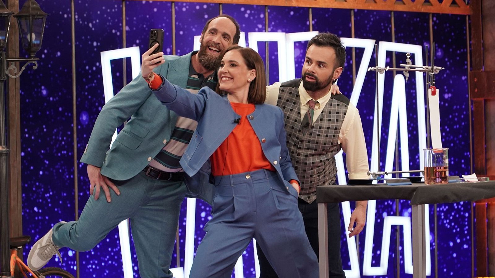 Goyo Jiménez, Raquel Martos y Ricardo Moure posan para una selfie en un plató de televisión con temática científica, luciendo atuendos variados y rodeados de elementos como un farol y equipo de laboratorio.