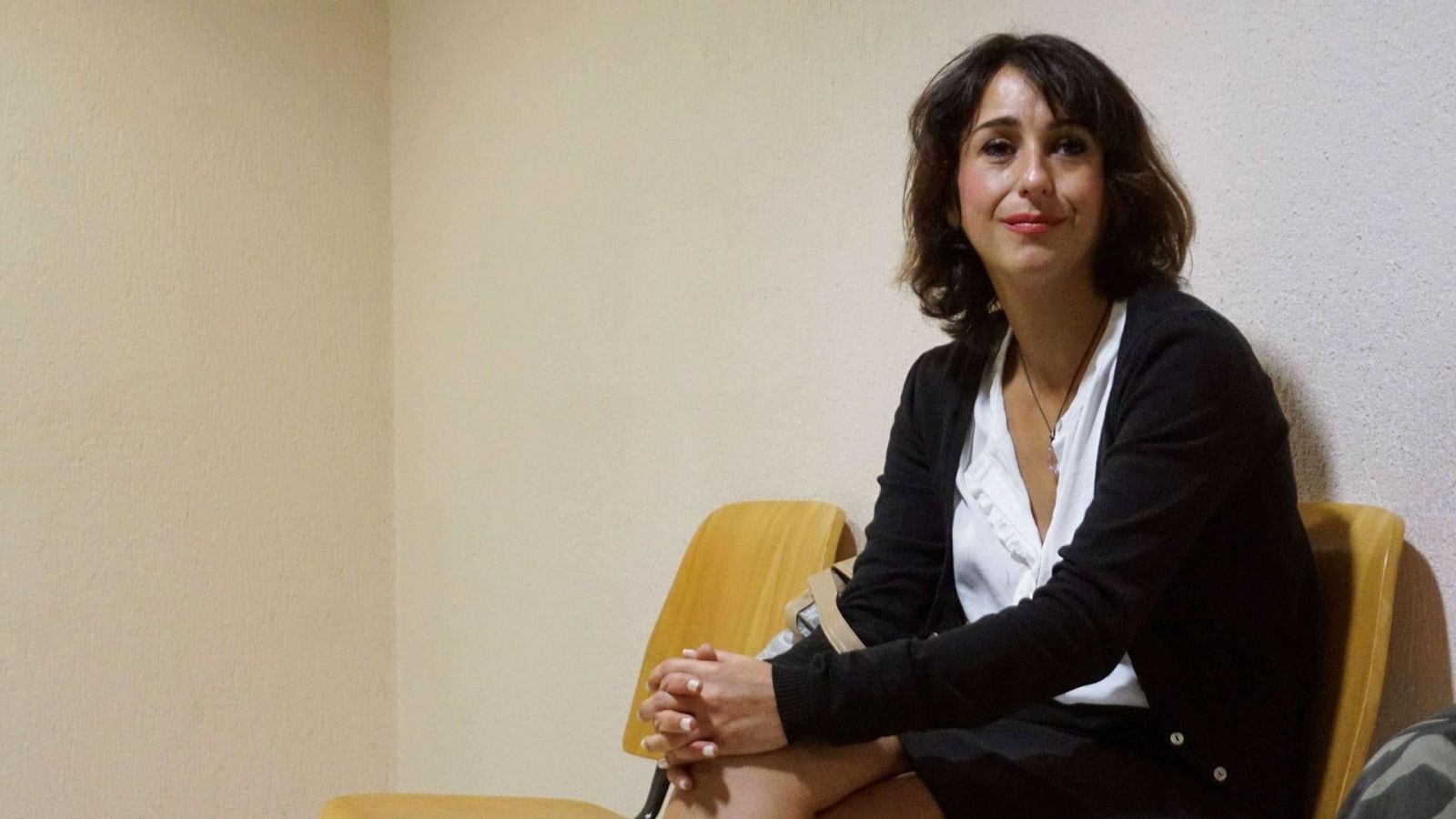 Mujer con blusa blanca y chaqueta negra sentada en sala de espera de paredes crema, con expresión serena y leve sonrisa.