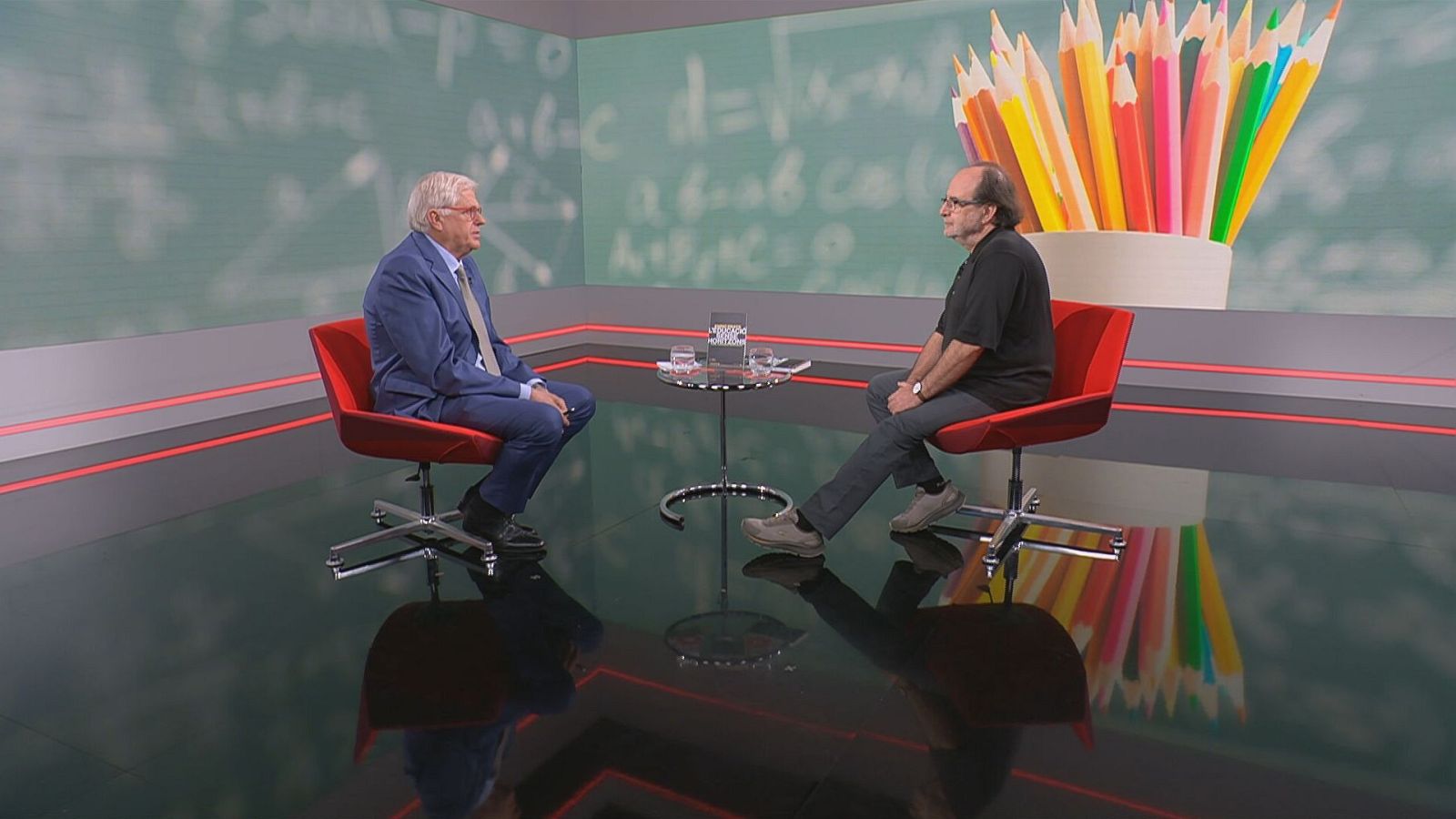 Lluís Falgàs entrevista Enric Prats en un estudi de televisió. Falgàs porta vestit blau, Prats samarreta negra. Al fons, una pissarra verda amb fórmules i llapis de colors.