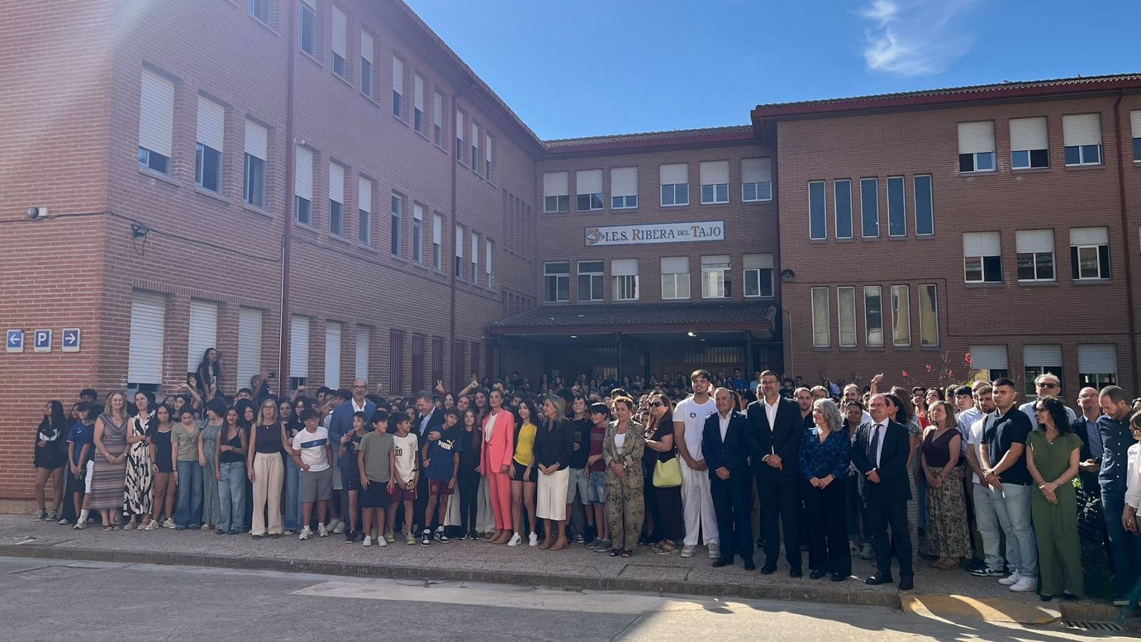 La Reina Letizia, vestida de coral, visita el I.E.S. Ribera del Tajo, rodeada de estudiantes y autoridades en un evento exterior.