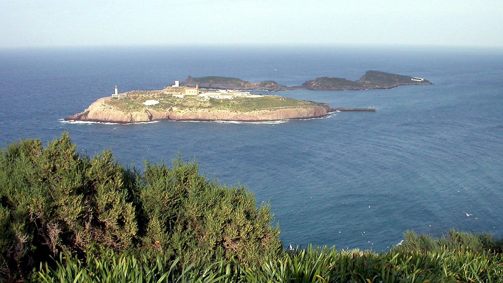 Archipiélago rocoso con faro blanco, edificios y muelle en la isla principal. Mar azul intenso y cielo despejado. Vegetación en primer plano.