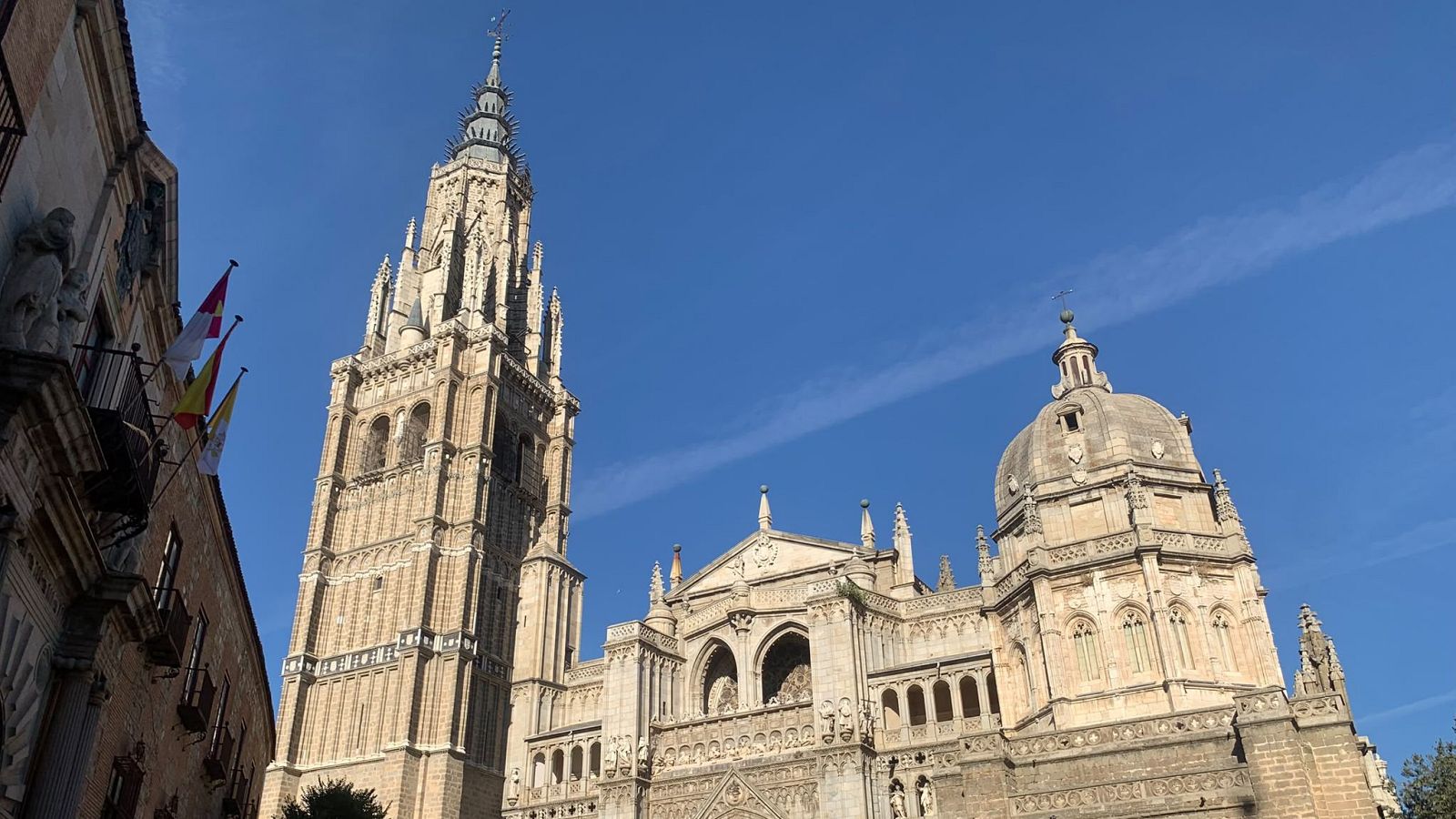 Vista ascendente de la Catedral de Toledo, con su campanario gótico y cúpula renacentista. En el balcón de un edificio cercano ondean las banderas de España y Toledo.