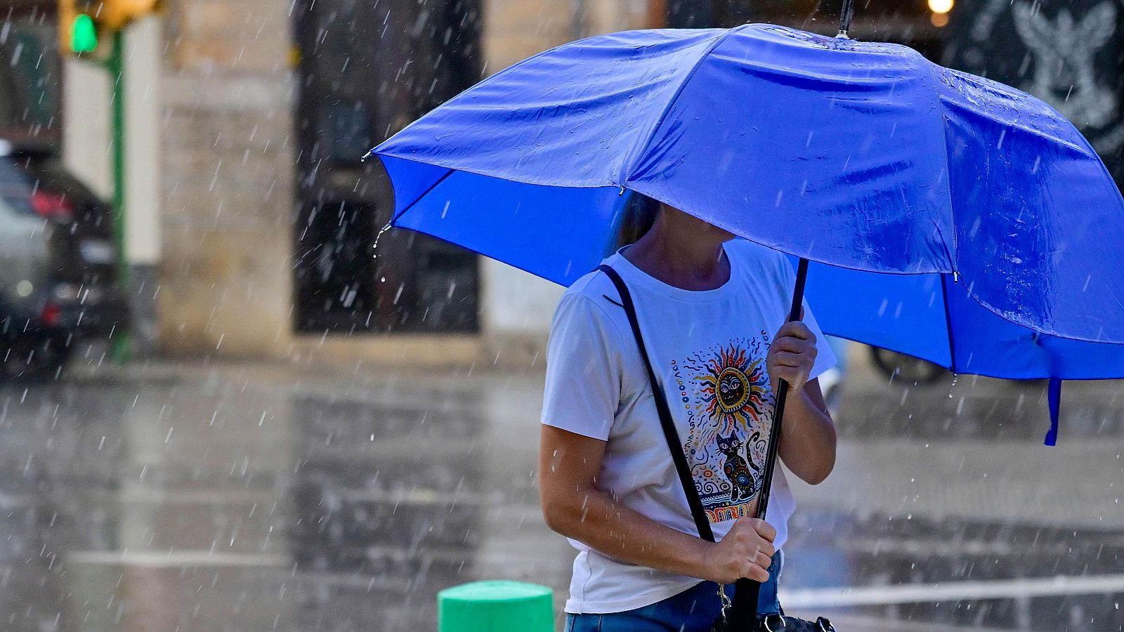 Persona con camiseta gráfica de sol y gato, protegida por paraguas azul bajo intensa lluvia urbana. Gotas y reflejos visibles.