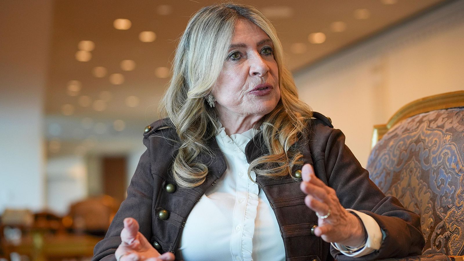 Ivonne Baki, candidata a la Secretaría General de la ONU, gesticula con las manos en una entrevista, luciendo una chaqueta militar y cabello rubio ceniza. Fondo desenfocado con luces cálidas.
