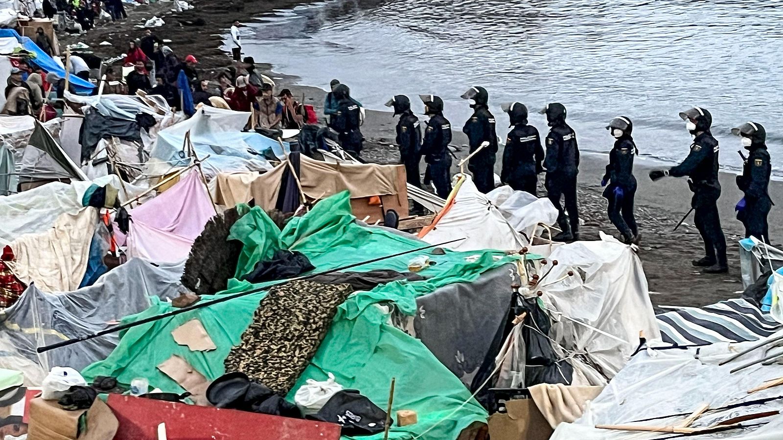 Campamento de migrantes en la playa de Benítez con presencia de fuerzas de seguridad. Refugios improvisados y personas dispersas en la orilla.