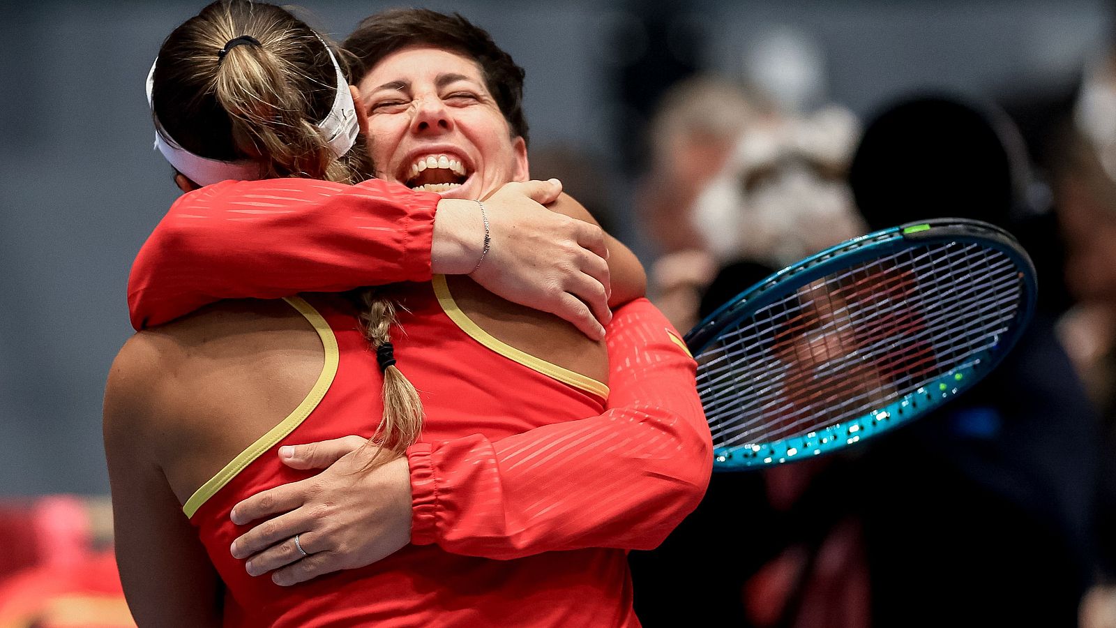 Carla Suárez, con los ojos cerrados y una gran sonrisa, abraza a una compañera de equipo. Ambas visten uniformes rojos con detalles amarillos, sugiriendo un momento de celebración en un evento deportivo.