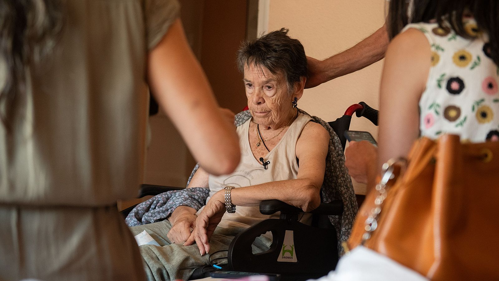 Primer plano de Maricarmen, mujer mayor en silla de ruedas, con expresión seria. Se aprecian otras personas y detalles de vestimenta y accesorios en un entorno de apoyo.