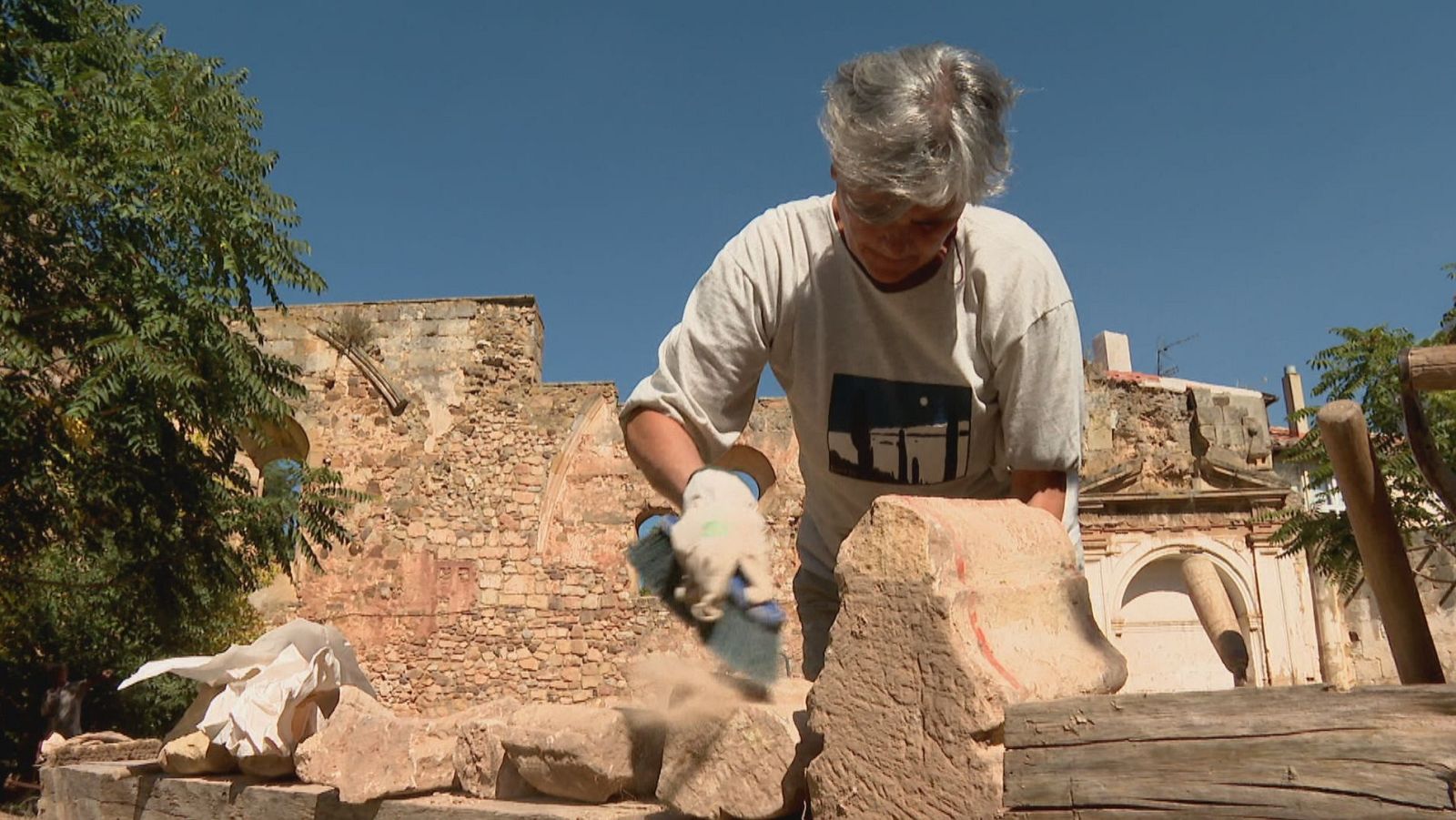Arqueóloga Helena Inglada, con guantes y herramienta, limpia una piedra en un sitio de excavación con ruinas antiguas y cielo azul.