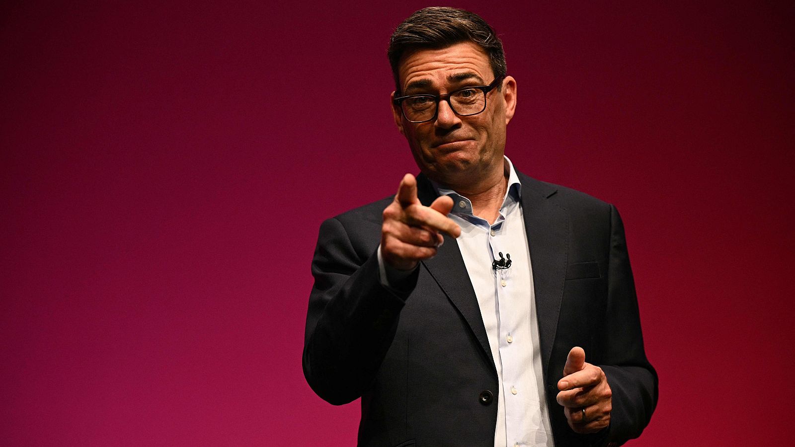 El político británico Andy Burnham, con gafas y traje sin corbata, sonríe y apunta con el dedo índice hacia la cámara sobre un fondo magenta.