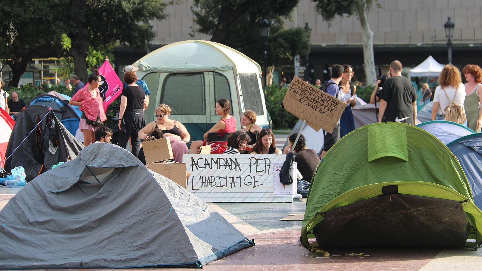 Protesta en una plaça amb tendes de campanya i cartells on es llegeix 'ACAMPADA PER L'HABITATGE' i 'WELCOME TO SPAIN TOURISTS, THIS IS THE REALITY OF THE COUNTRY' amb gent reunida.