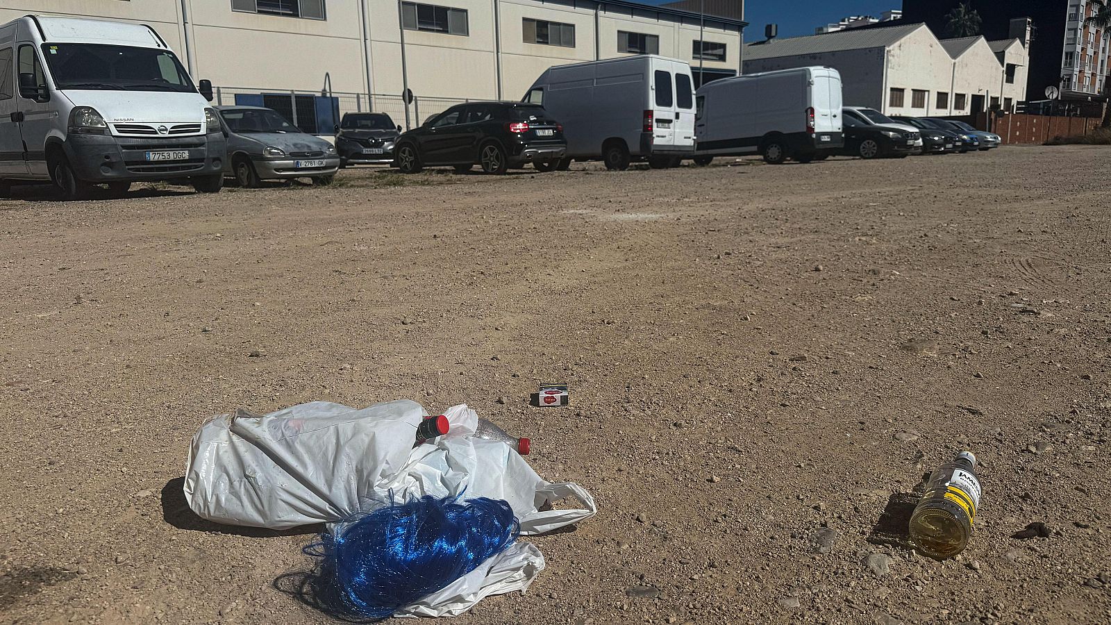 Descampado de tierra con residuos como botellas y bolsas plásticas en primer plano, y vehículos aparcados frente a naves industriales al fondo bajo un cielo despejado.
