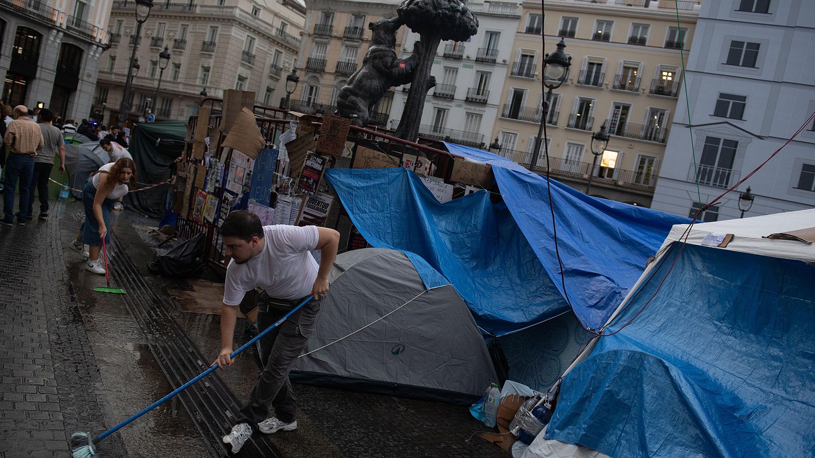 Multitud de manifestantes marcha por una calle céntrica con edificios clásicos, liderados por un hombre con barba que grita consignas.