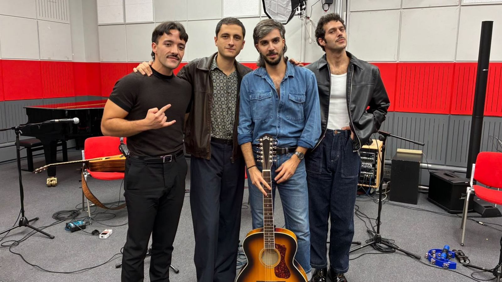 Cuatro músicos posan en un estudio de radio con paneles acústicos blancos y rojos, destacando una guitarra de doce cuerdas y un piano de cola al fondo.