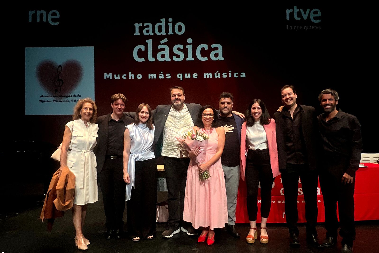 Grupo de nueve personas posando sonrientes sobre un escenario teatral bajo iluminación cálida frente a un fondo oscuro con logotipos de Radio Clásica.