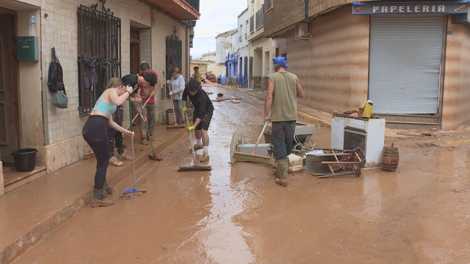 Vecinos y jóvenes limpian con escobas y cepillos una calle estrecha cubierta de lodo y muebles destrozados tras una riada.