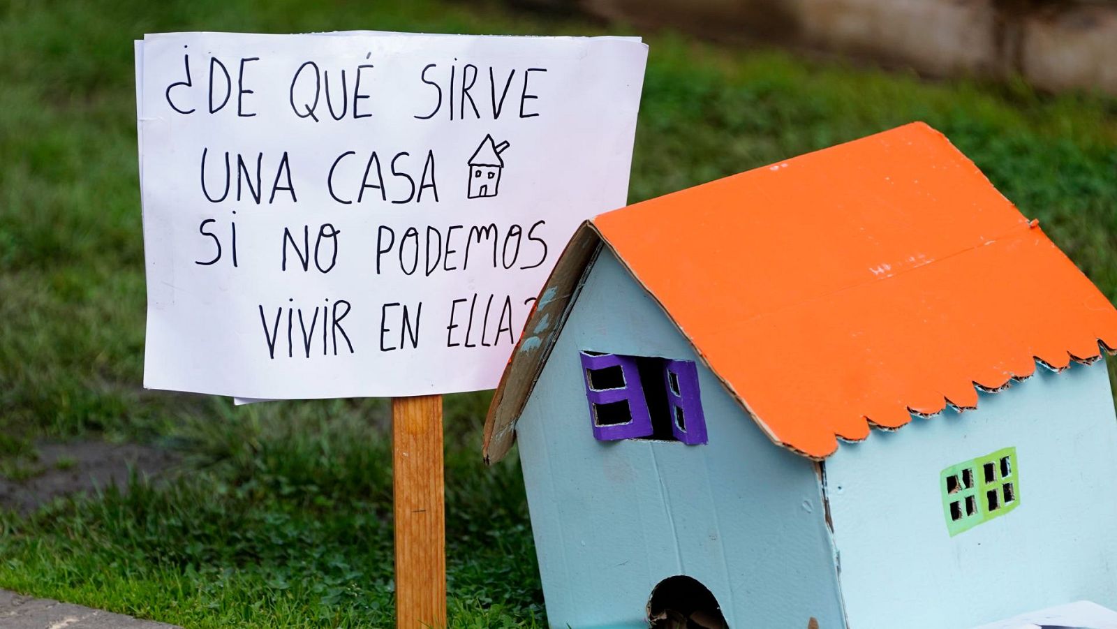 Cartel reivindicativo y maqueta artesanal de casa de cartón sobre césped en protesta por el acceso a la vivienda.