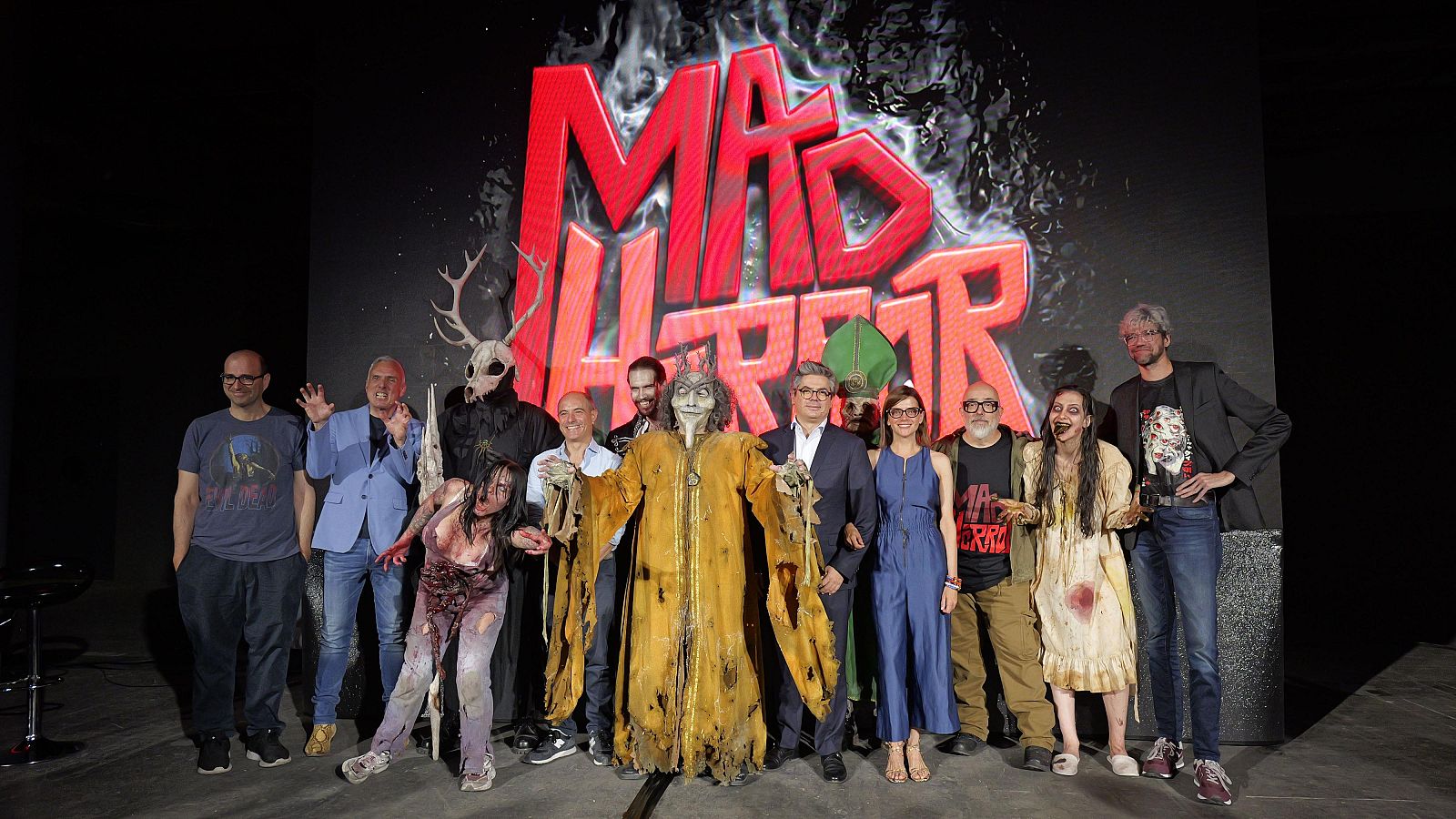 Grupo de personas e invitados caracterizados como criaturas de terror posan frente a un gran logotipo rojo de 'MAD HORROR' en un escenario.