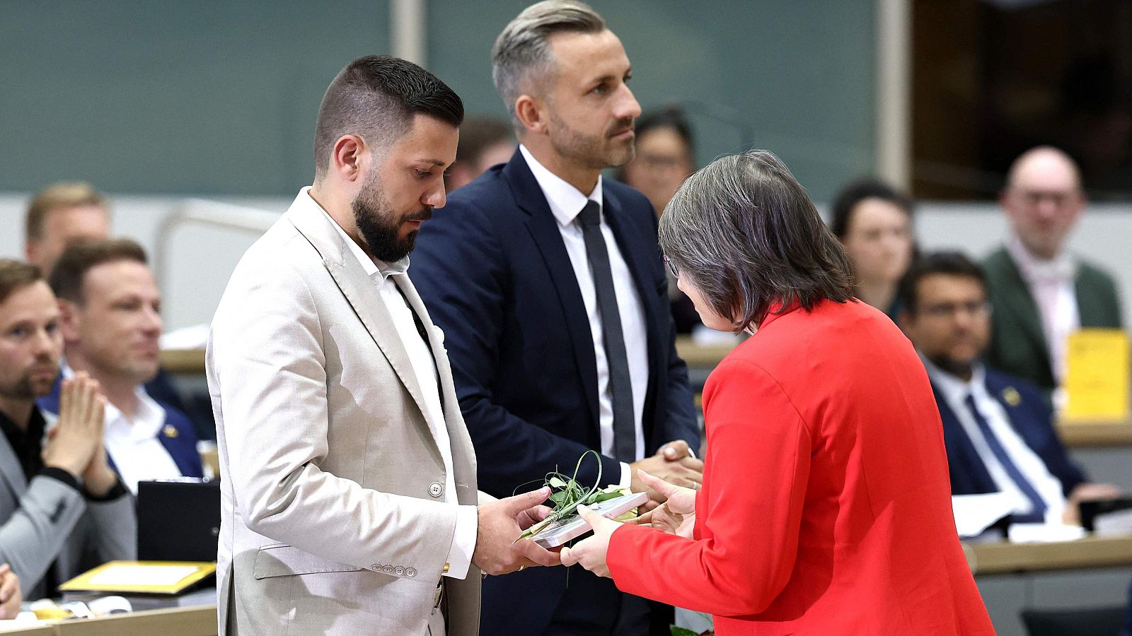 Diputada con chaqueta roja entrega una constitución y una flor a un diputado con traje beige ante la atenta mirada de otro compañero en el parlamento.
