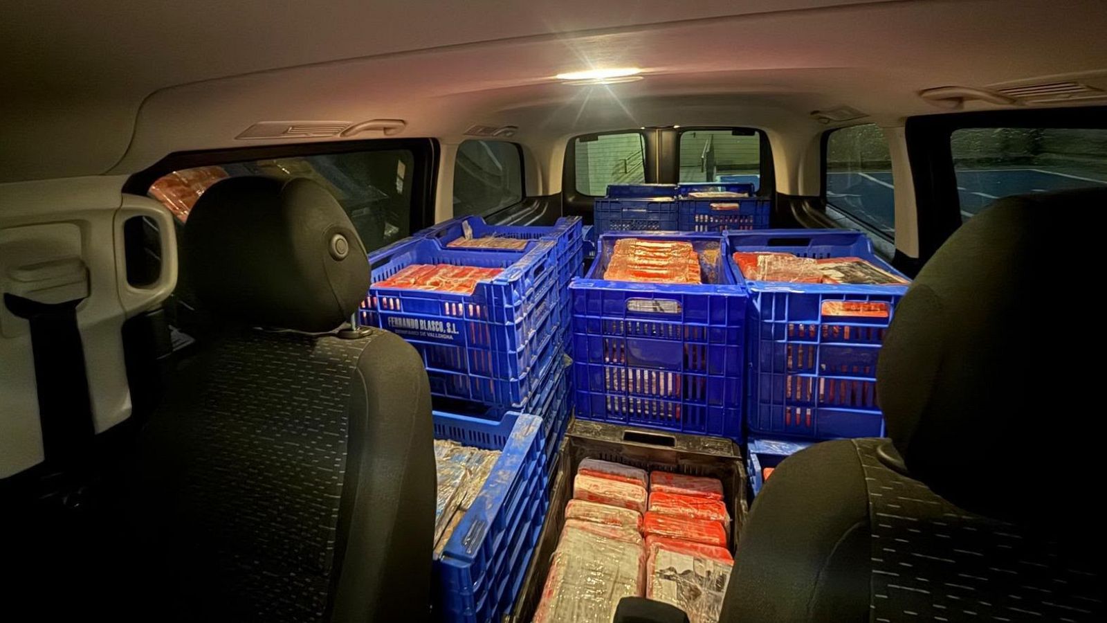 Interior de una furgoneta con el espacio trasero abarrotado de cajas azules llenas de paquetes envueltos en plástico de colores.