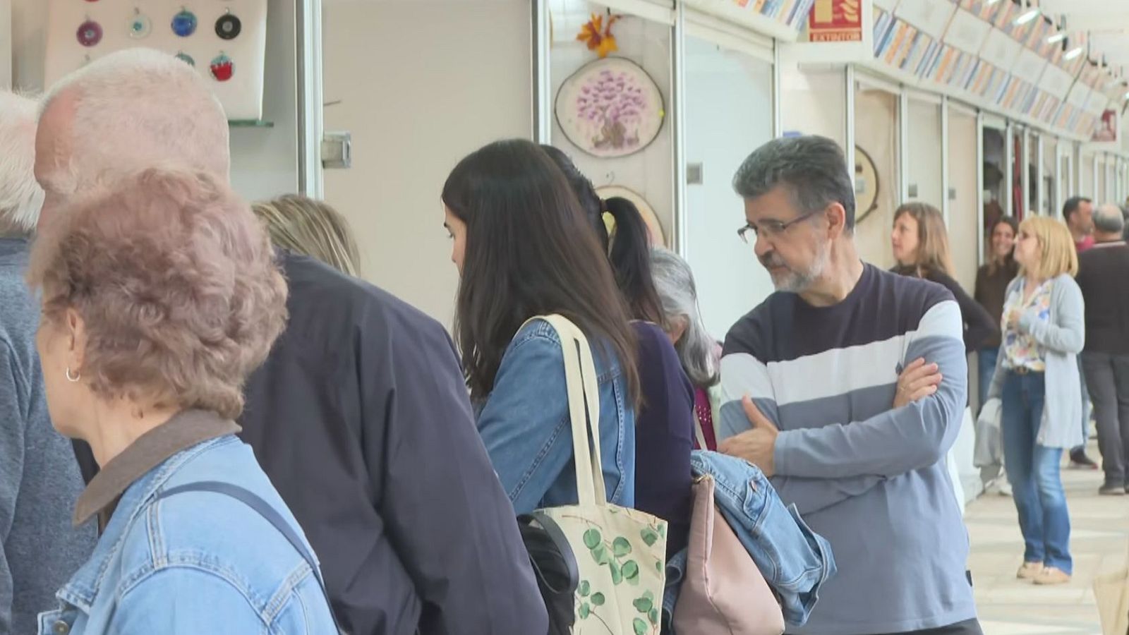 Concurrido pasillo interior de una feria de artesanía con casetas blancas y visitantes observando los puestos.