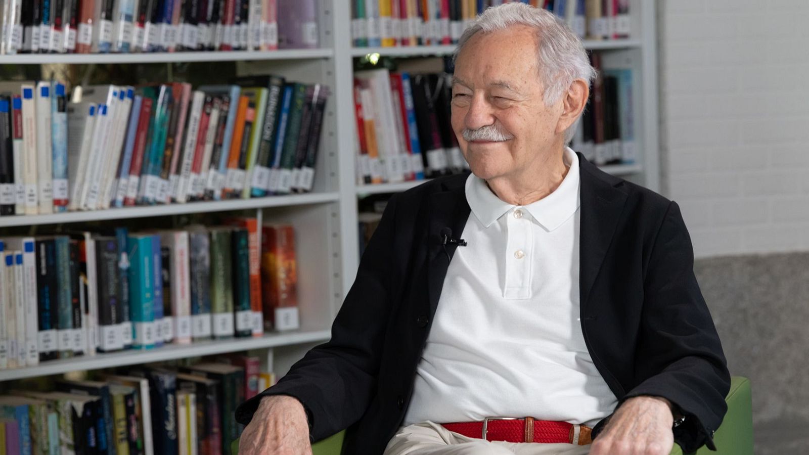 El escritor Eduardo Mendoza sonríe sentado en una butaca, con chaqueta negra y micrófono de solapa, frente a una estantería de libros.