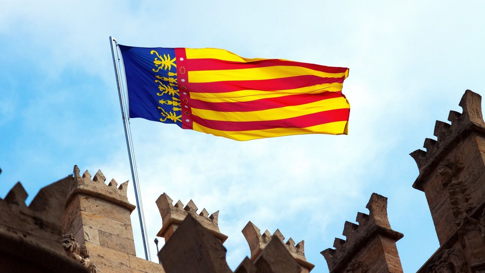 La Real Senyera de la Comunidad Valenciana ondea sobre las almenas de piedra de las Torres de Serranos bajo un cielo azul.