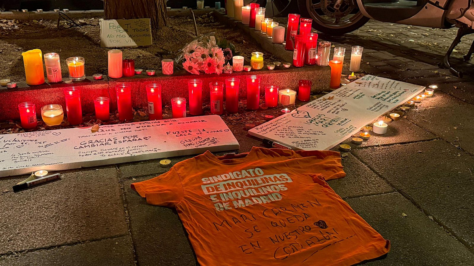 Memorial nocturno en la calle con velas encendidas, carteles manuscritos de homenaje, un ramo de flores y una camiseta naranja colocados sobre la acera.