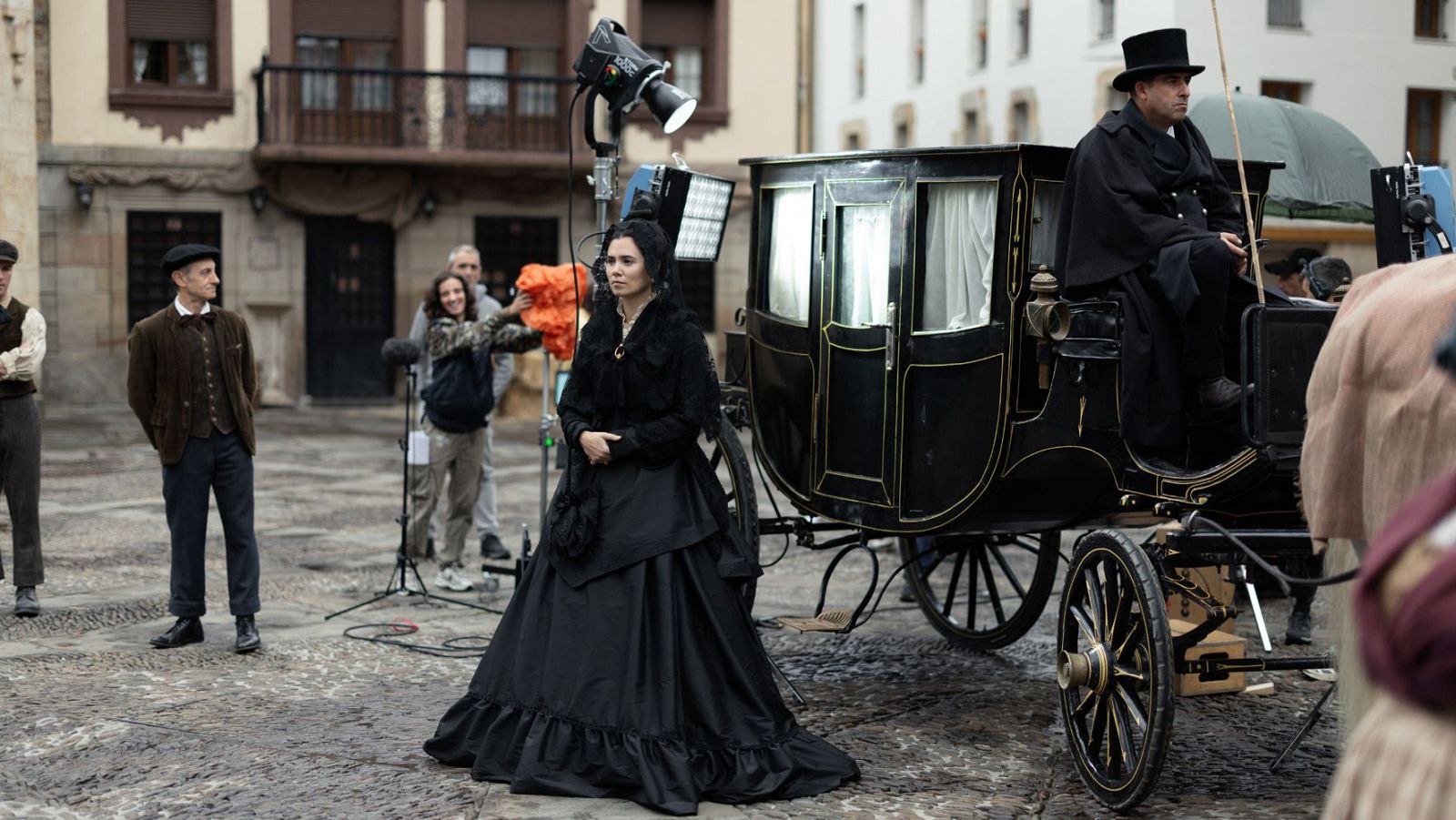 Imagen de un rodaje histórico con actores vestidos de época junto a un carruaje y personal técnico con equipamiento actual en una plaza empedrada.