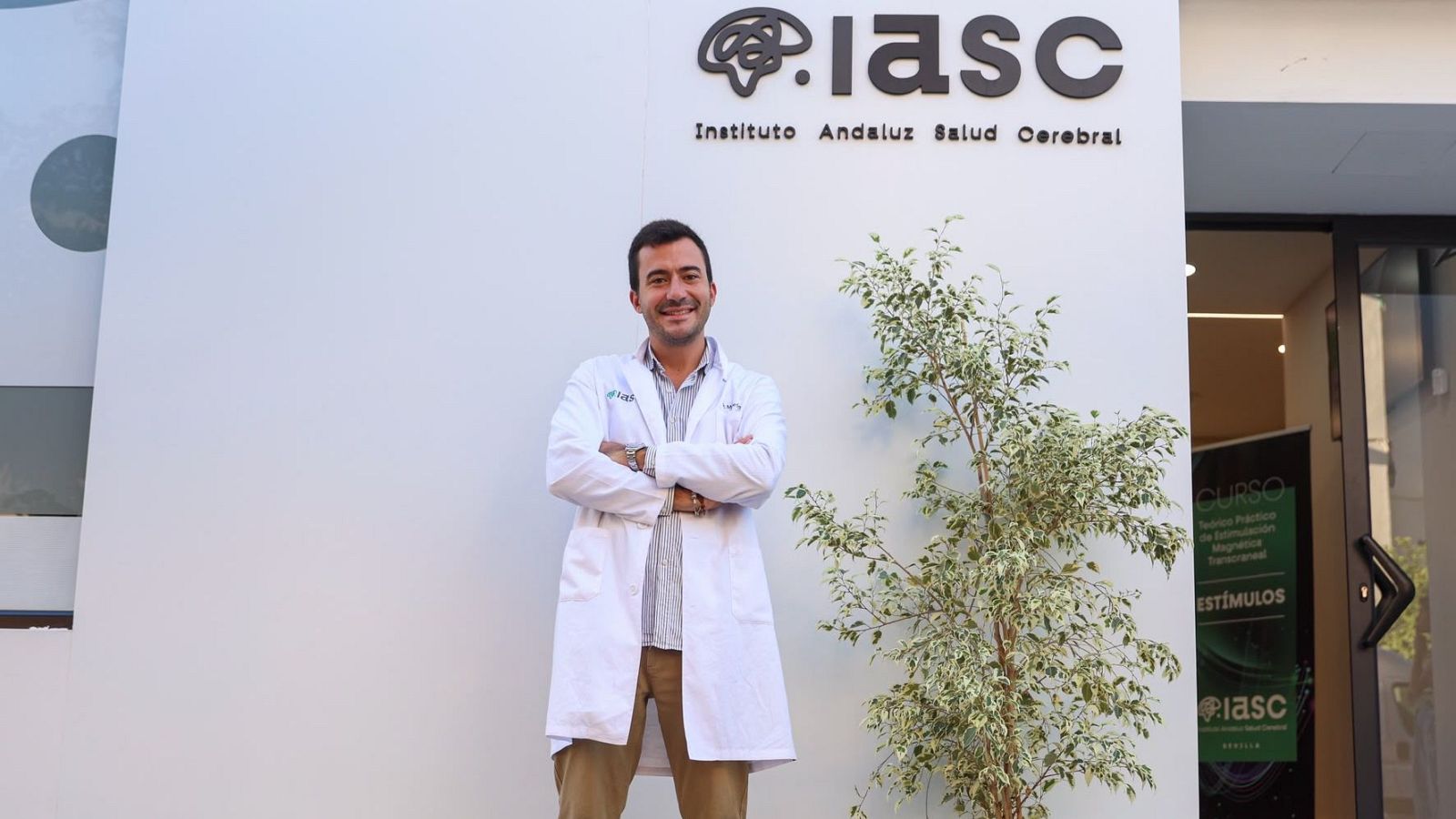 El psiquiatra Álvaro Moleón posa sonriente con bata blanca frente al logotipo del Instituto Andaluz de Salud Cerebral.
