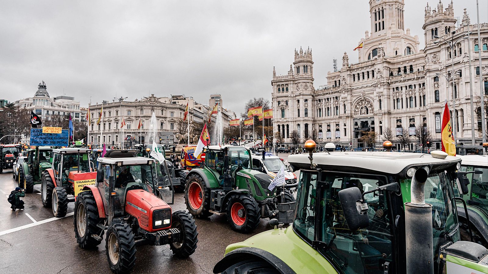Una protesta de agricultores en Madrid muestra tractores y pancartas en una calle, con el Palacio de Cibeles visible al fondo, en un día con el cielo cubierto.