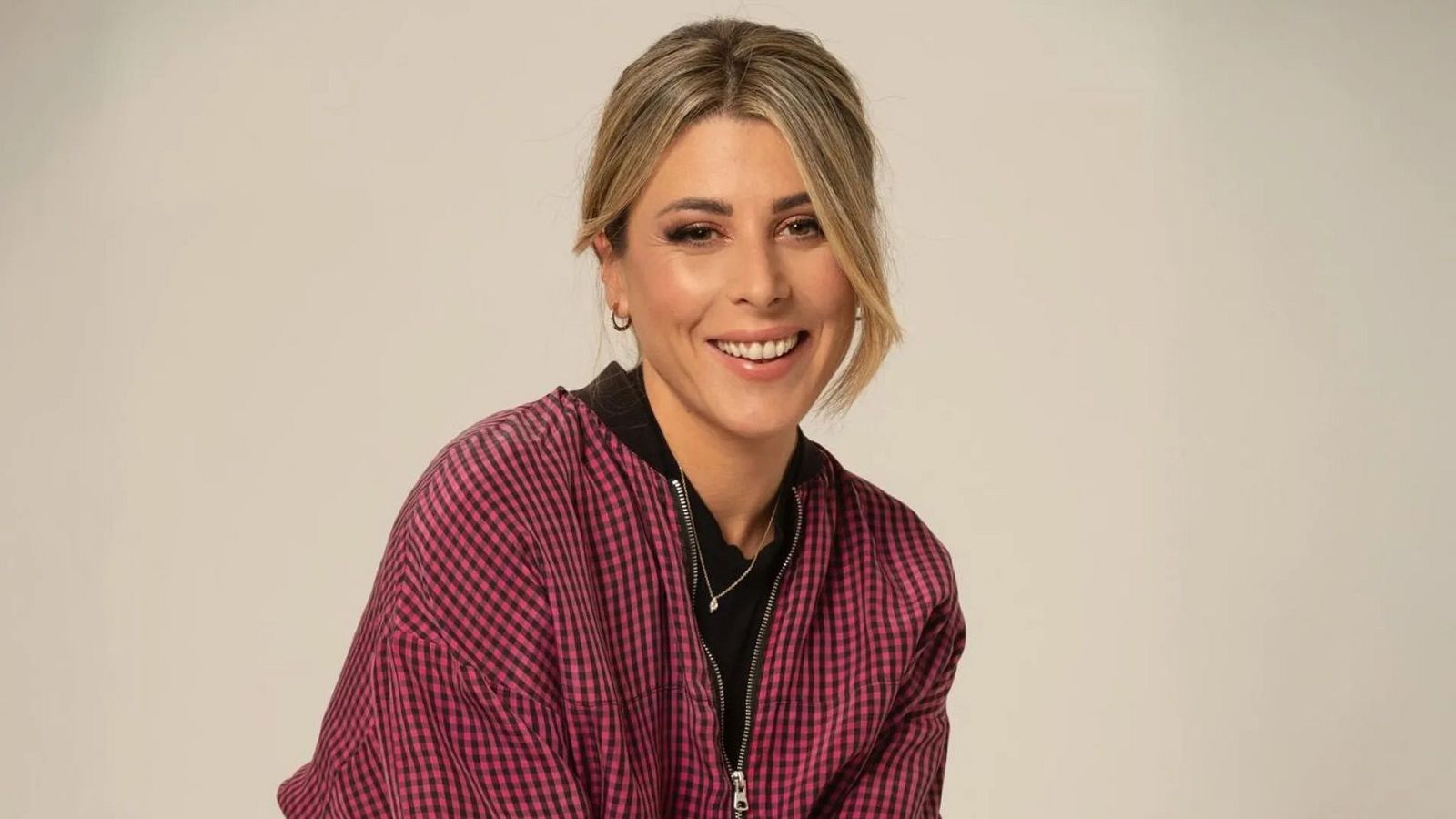 La cómica Valeria Ros posa sonriente en un estudio con fondo beige, luciendo una chaqueta de cuadros rojizos y negros.