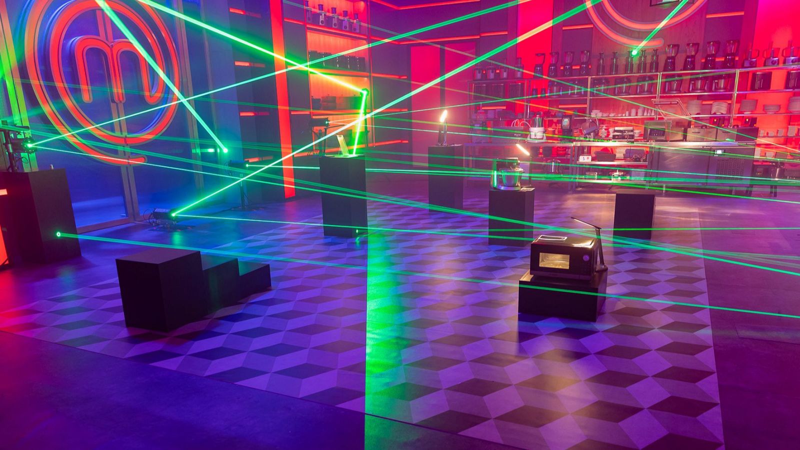 Plató de televisión iluminado en tonos rojos y fucsias con una densa red de láseres verdes y pedestales con electrodomésticos.