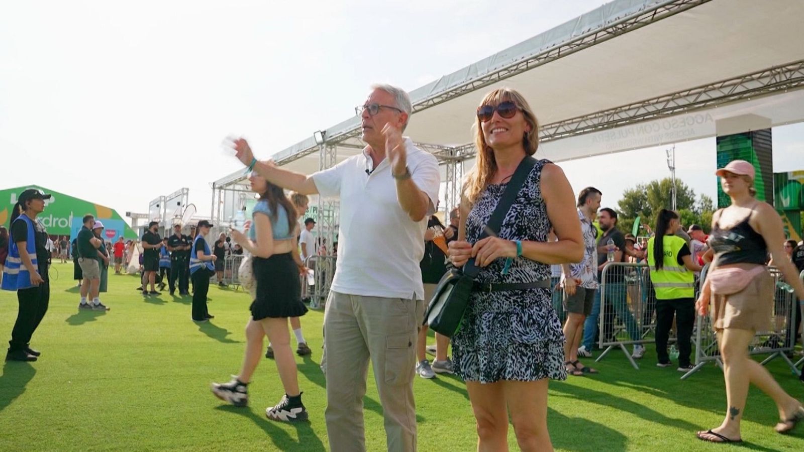 Un reporter amb polo blanc i micròfon de solapa gesticula davant la càmera en un festival de música a l'aire lliure.