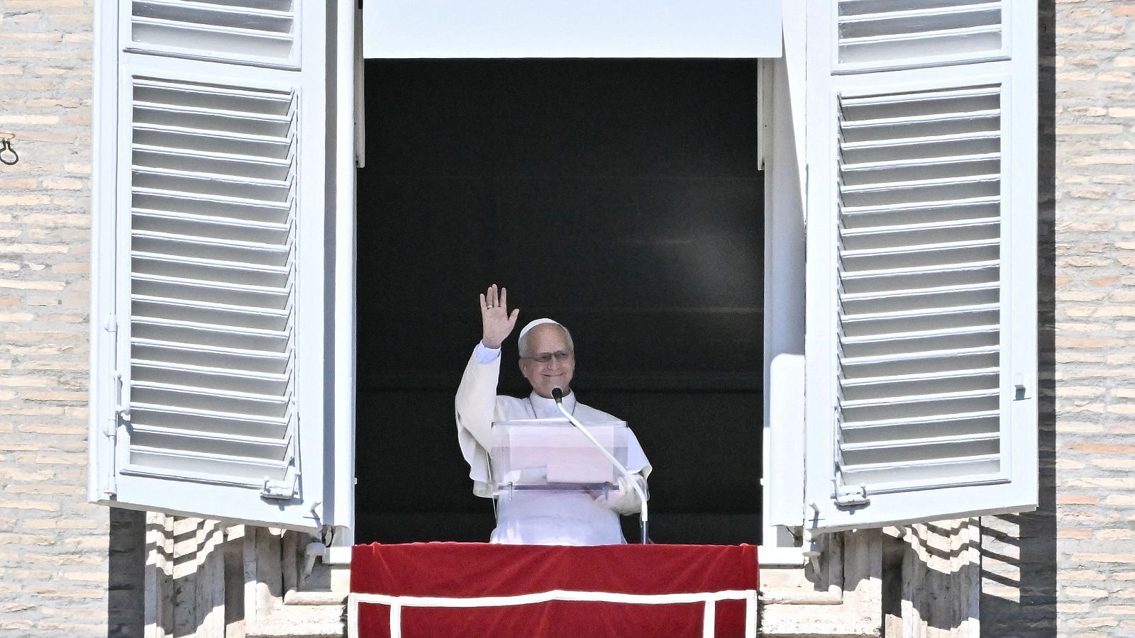 Un pontífice con sotana blanca y solideo sonríe y saluda con la mano alzada desde la ventana del Palacio Apostólico.