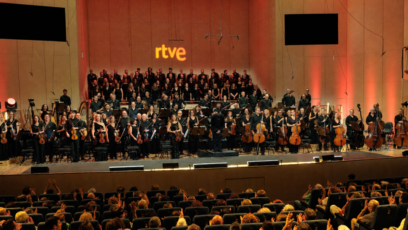 Vista desde la audiencia de un auditorio donde el público aplaude a la Orquesta y Coro de RTVE sobre el escenario iluminado con tonos cálidos.