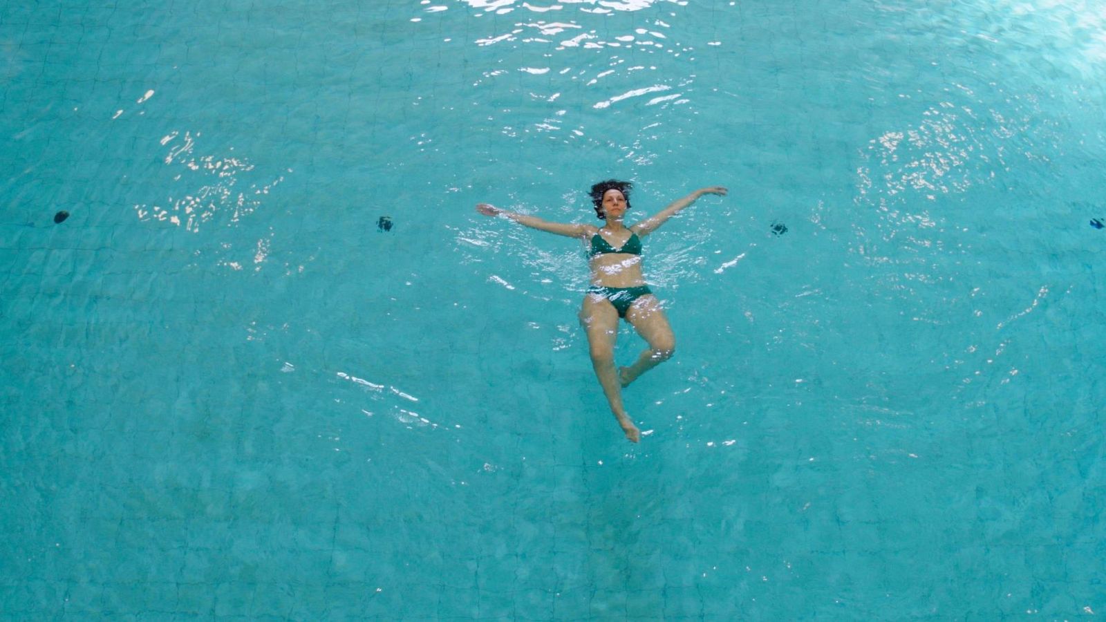 Mujer flotando relajada boca arriba en el centro de una piscina de aguas turquesas con bikini verde.
