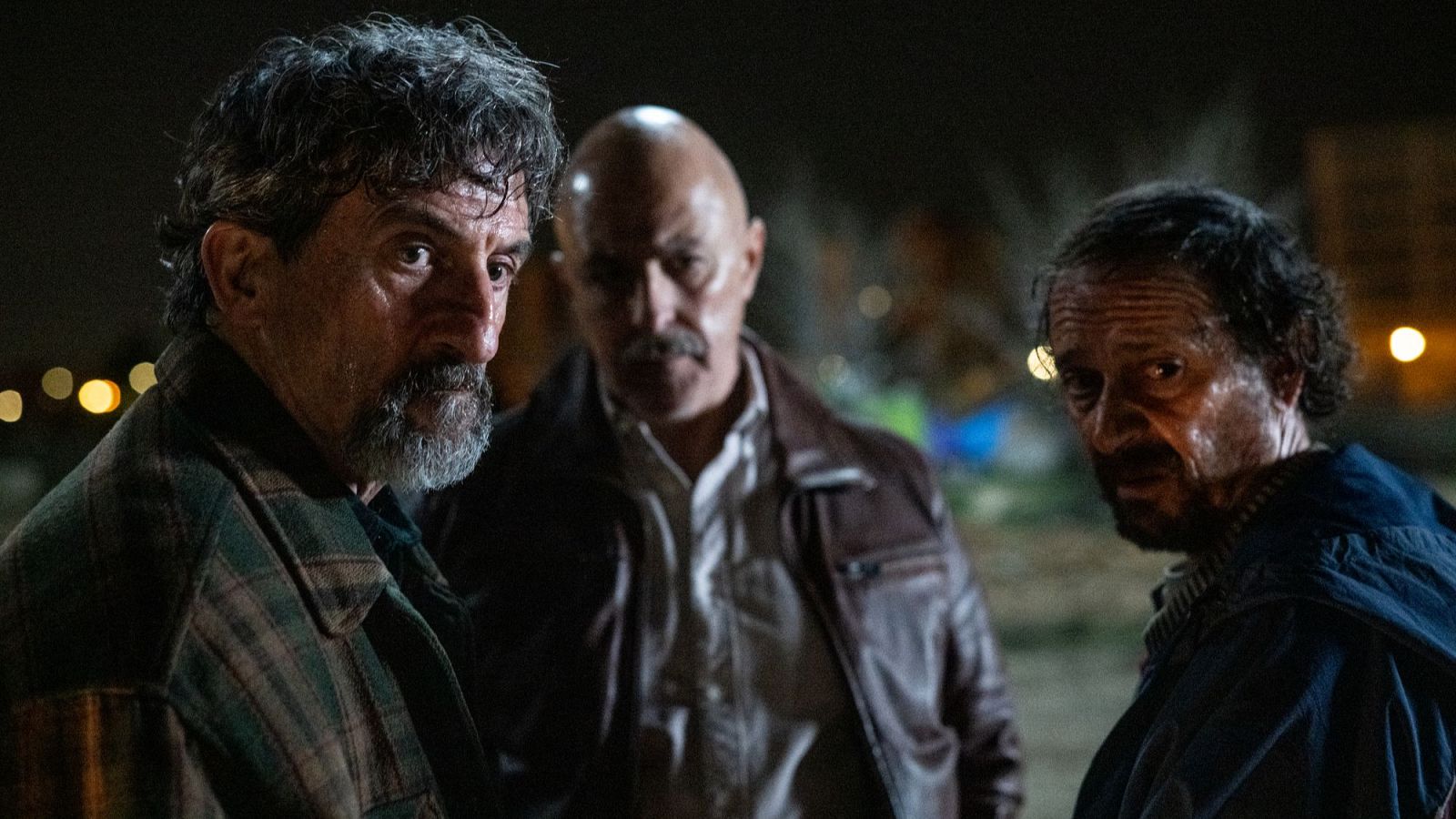 Plano medio nocturno de tres hombres con expresiones serias y tensas en un entorno urbano oscuro y desenfocado con luces lejanas.