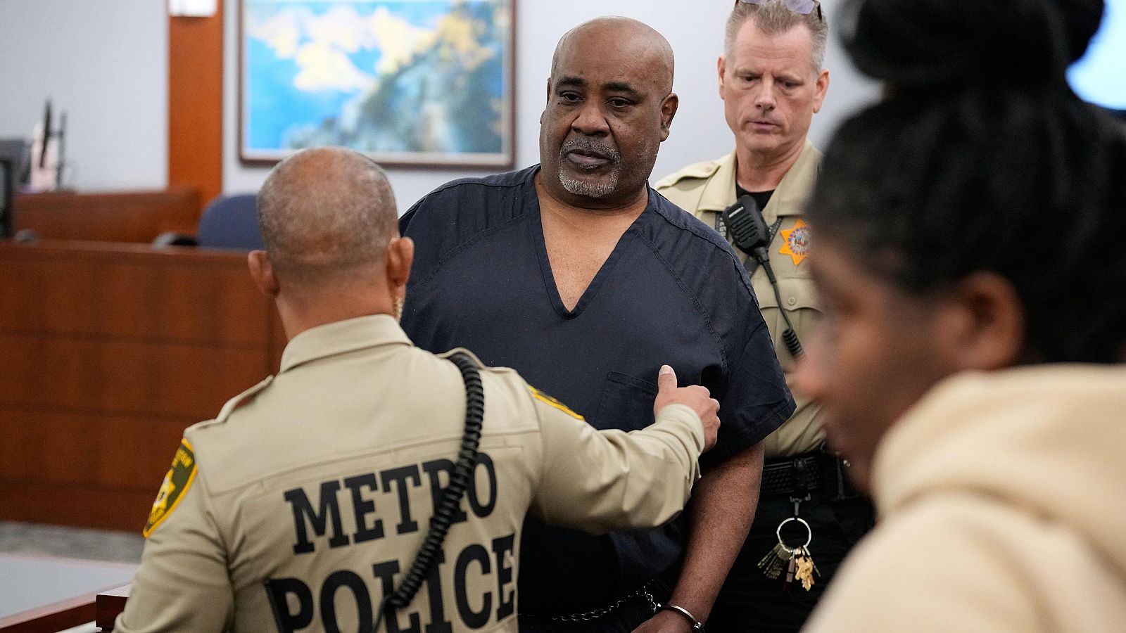 Duane Davis, con uniforme penitenciario y esposado, es custodiado por agentes de policía en una sala judicial.