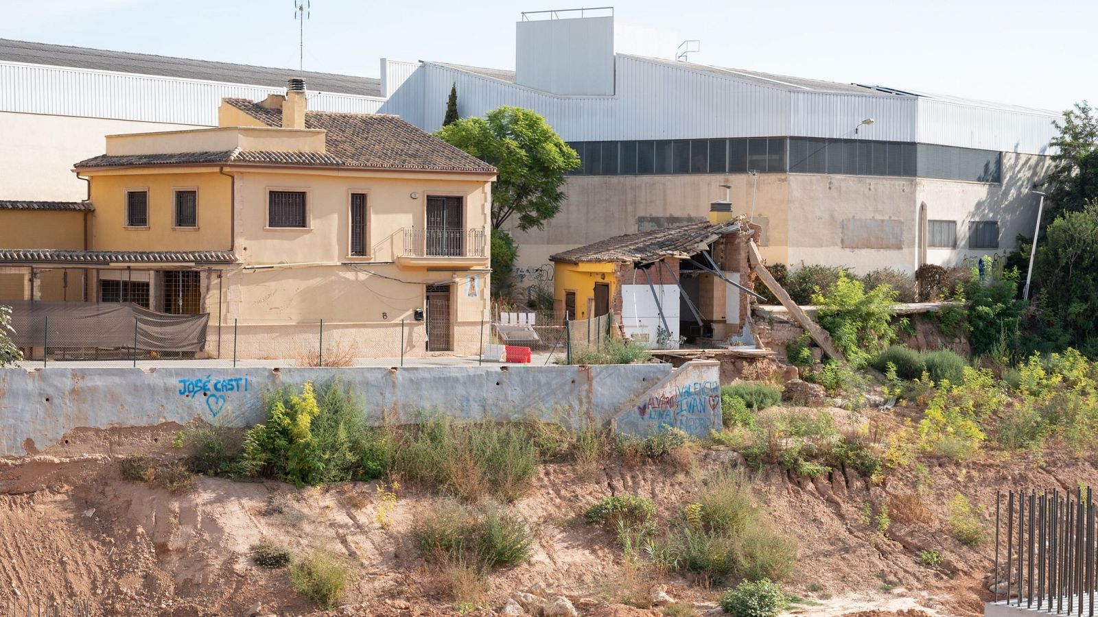 Dos casas, una en buen estado y otra en ruinas, se alzan sobre un terreno elevado con un muro de contención con grafitis. El contraste visual sugiere desigualdad y precariedad en el entorno.