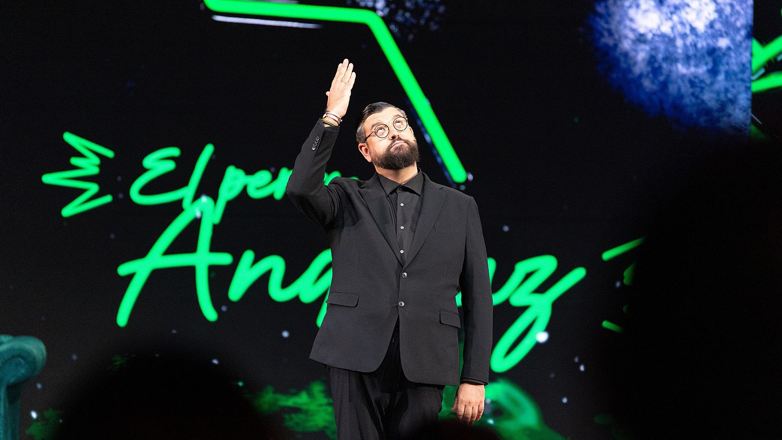 Manu Sánchez gesticula en el escenario con traje negro y gafas, frente a una pantalla iluminada en verde neón.