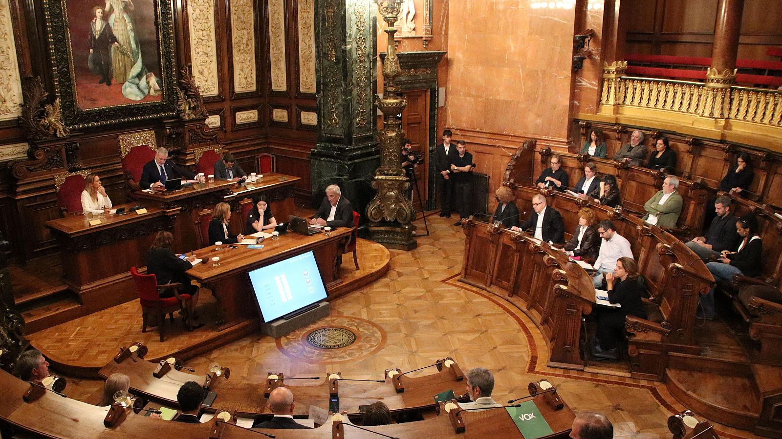Vista en picat del ple municipal de l'Ajuntament de Barcelona amb regidors asseguts a les bancades de fusta i la presidència al fons.