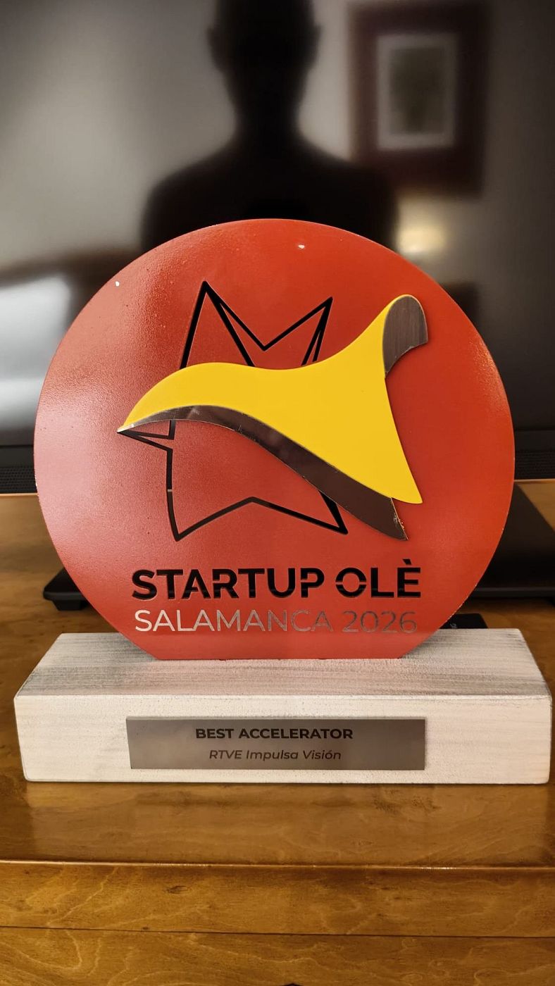 Primer plano de un trofeo institucional con pieza ovalada roja y base de madera que incluye una placa con el texto «BEST ACCELERATOR» y «RTVE Impulsa Visión».
