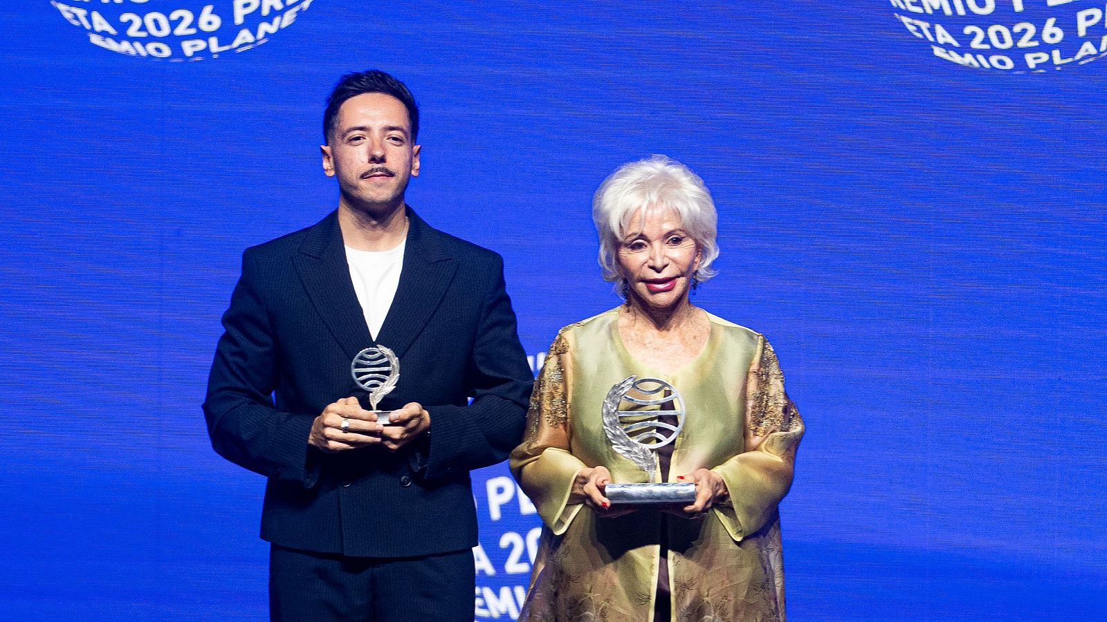 Dos autores posan sonrientes en un escenario con sus estatuillas plateadas del Premio Planeta frente a un fondo azul con letras blancas.
