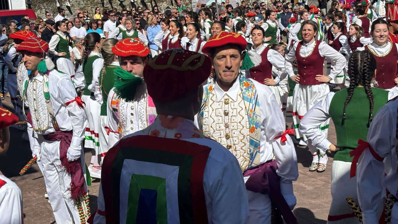 Grupo de danzantes con trajes tradicionales de bolantes, destacando pecheras bordadas y gorros rojos con borlas, ante una multitud en un día soleado.
