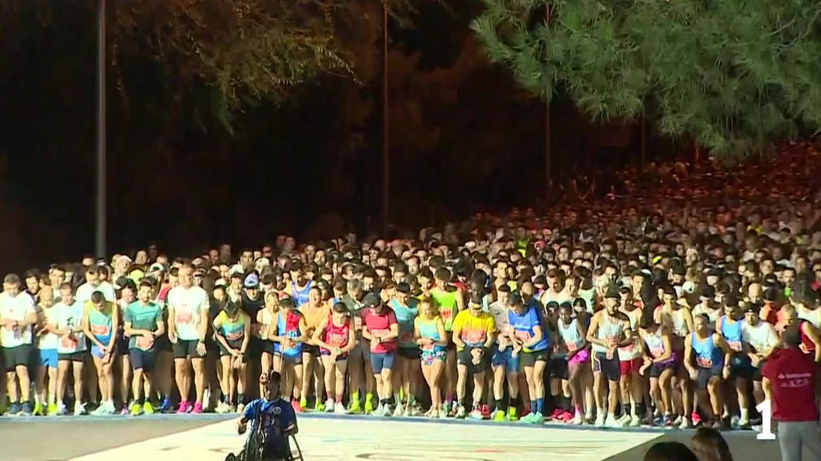 Atleta en silla de ruedas en la línea de salida de una multitudinaria carrera atlética nocturna iluminada por focos.