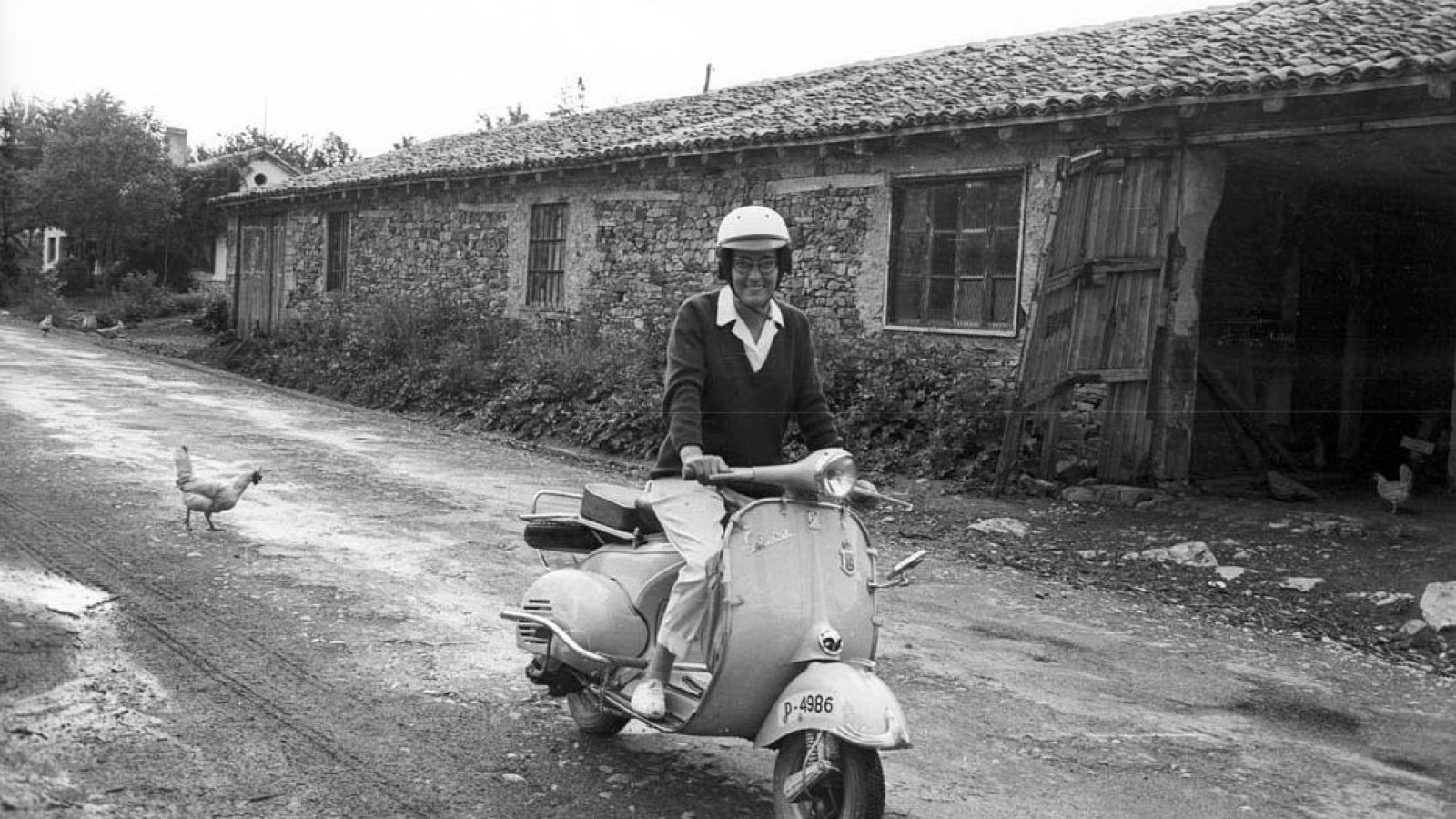 Fotografía en blanco y negro de Miguel Delibes sonriendo sobre una Vespa en un camino rural junto a casas de piedra.