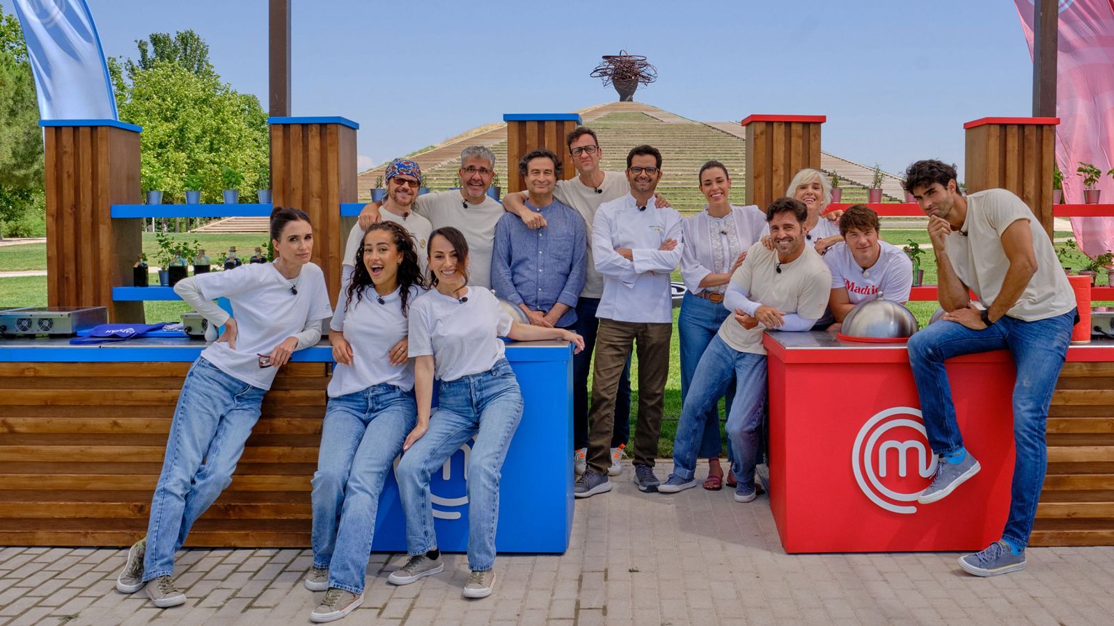 Participantes y jueces de un programa culinario posan sonrientes en un parque soleado junto a puestos de cocina móviles.