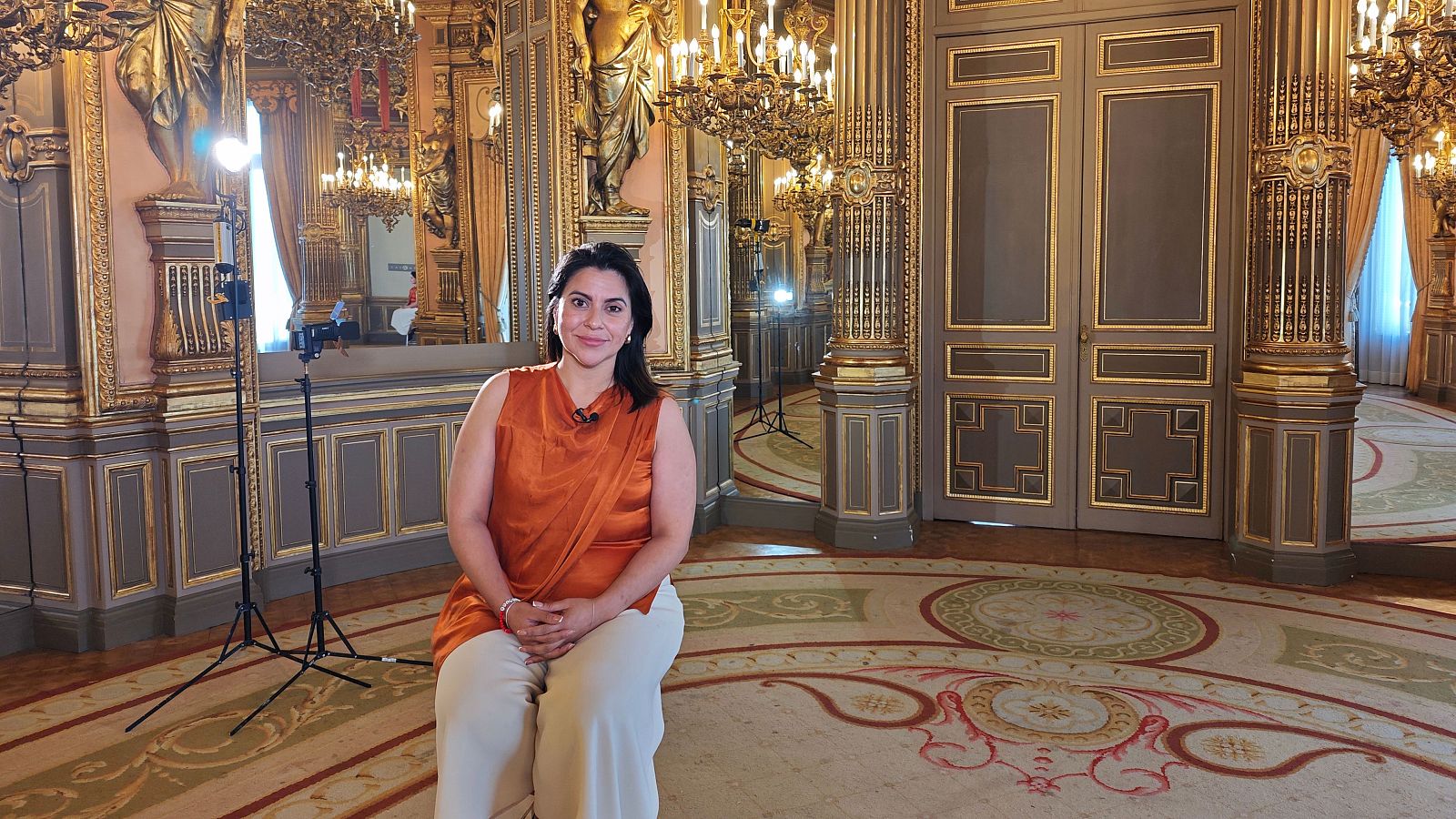 La activista Olimpia Coral Melo sonríe sentada en un elegante salón neoclásico con columnas doradas y espejos durante una entrevista.