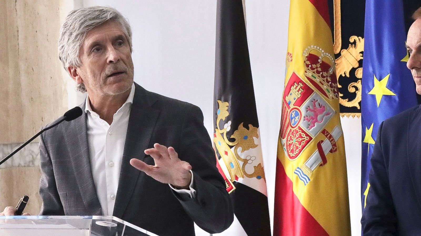 Plano medio del ministro Fernando Grande-Marlaska hablando ante un micrófono con traje gris y camisa blanca, frente a las banderas de Ceuta, España y la Unión Europea.