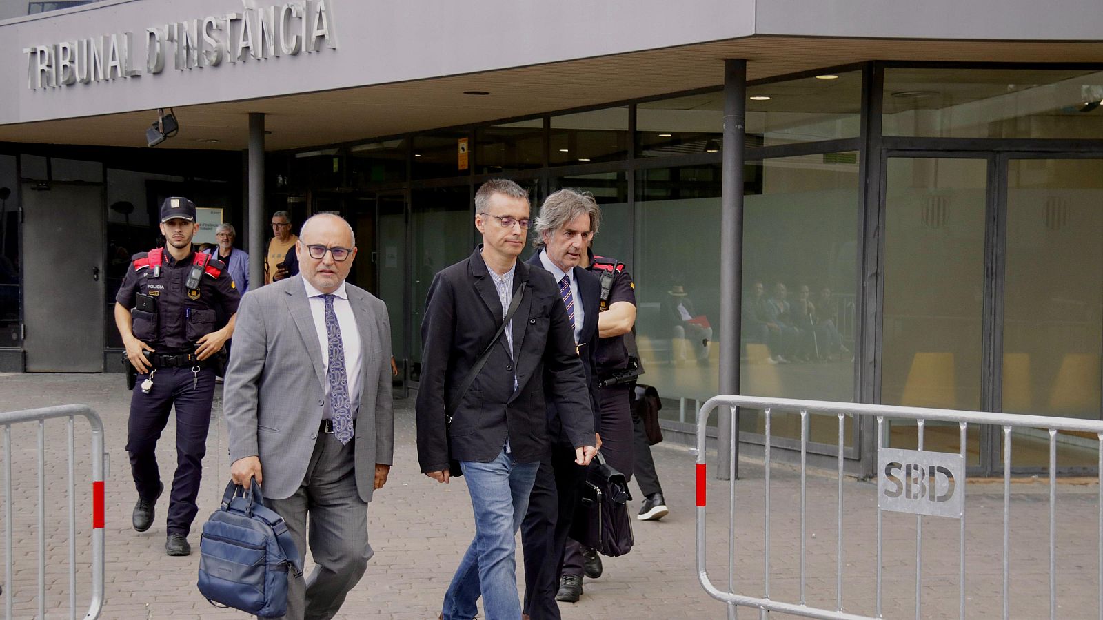 Santi Laiglesia camina serio a la salida de los juzgados junto a su equipo legal, luciendo chaqueta negra y vaqueros ante la fachada acristalada del edificio judicial.
